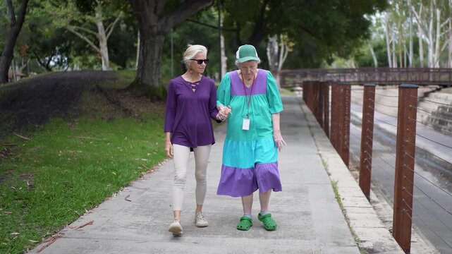 Daughter visits her mother in a park for a walk holding hands together, talking and enjoying their time while smiling and sharing memories.