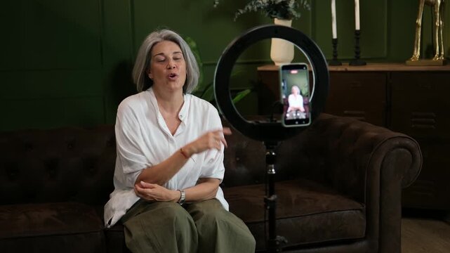 Elegant older woman speaking in front of a smartphone on a tripod with a ring light at home
