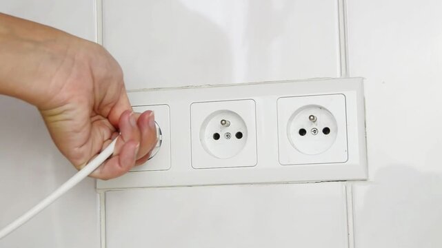 Close-up of a female hand plugging an electrical power plug into a wall socket. Everyday household action showing connecting an electronic device to the power supply, home electricity.