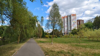 A river flows along a paved path in the park, and willows and other deciduous trees grow on grassy lawns. Behind them stands a brightly painted high-rise building. Sunny autumn weather and blue sky © Balser