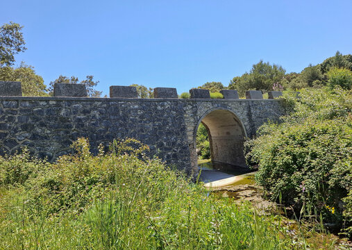 Ancient stone arch bridge spanning a river in a green area under a bright blue sky