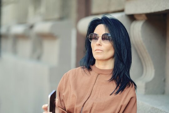 A woman wearing stylish sunglasses stands next to a historic building, casually holding her smartphone