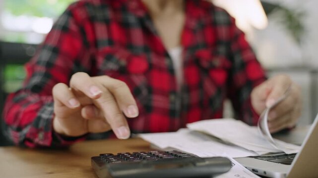 Closeup to hand of female accountant sat at desk with laptop computer using calculator to calculate company bills including wages utilities and taxes in office. Businesswoman working in her workplace.