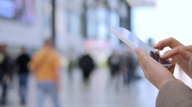 Hands hold smartphone in busy indoor public space. Person interacts with device while crowd moves behind. Focus stays on hands and screen. Ideal for digital connectivity, urban lifestyle