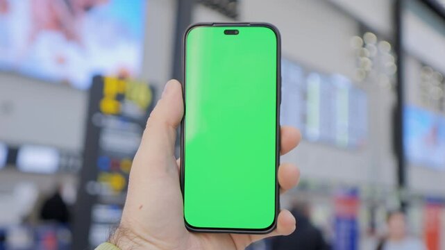 Hand holds phone with green screen. Background shows busy public space. Ideal for app previews or digital content. Blurred crowd suggests anonymous usage. Useful for tech presentations or UI design