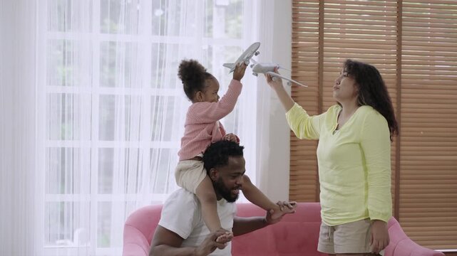 Happy African family. A cute daughter rides on her father's shoulders, holding and playing with a plane model with her mother, having fun and cheerful together on the sofa in the living room at home. 