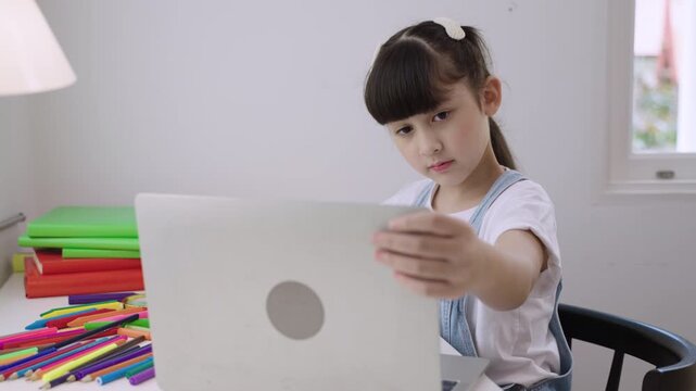 Cute Asian girl student sitting at desk using a laptop computer for studying online learning via video calling with remote teacher in a home school. Homeschooling education and new normal concept. 