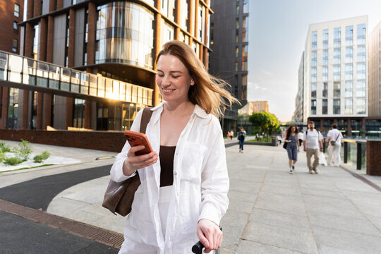 A woman checks her phone on a modern city sidewalk at golden hour