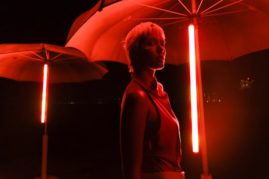 Model Stands Under Red Lights at Night Near Umbrellas