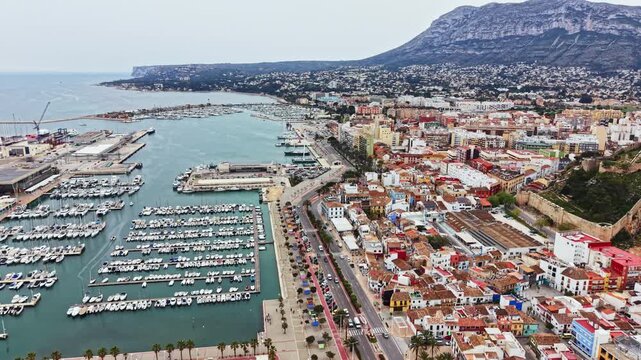 A view of a busy harbor filled with boats. A town with colorful buildings is located near the water and the base of a large mountain. The scene is lively and shows daily life by the sea.