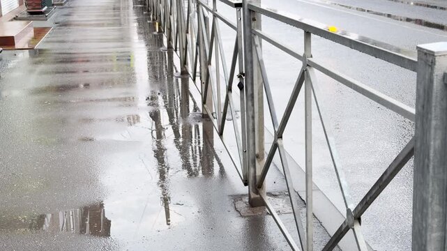 A metal safety railing stands alongside a rain-slicked city sidewalk. The large puddles cast clear, gloomy reflections, capturing a quiet and melancholic mood after a heavy downpour.