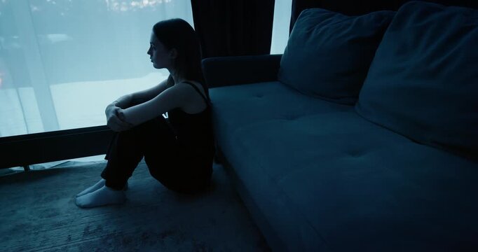 Alone woman sit floor by window at night. Sad young person looking out in dark room at home. Gloomy atmosphere, loneliness, depression, solitude. Melancholy mood, introspection, psychological struggle