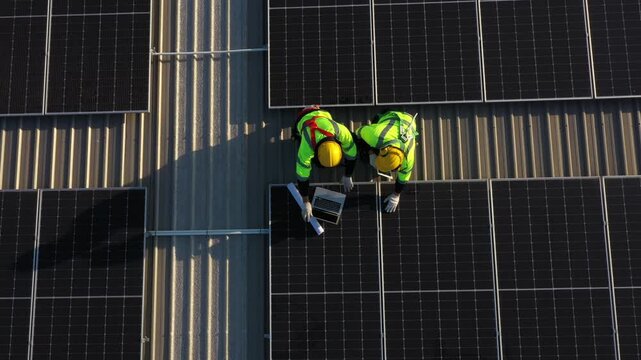 Renewable energy installation underway: Skilled workers mount solar panels on dark rooftop under bright sunlight, wearing yellow and green high-visibility vests and hard hats for safe operation.