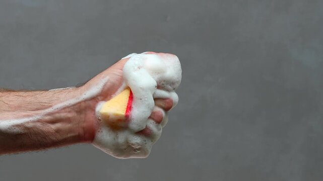 A man's hand in fist squeezes red washing sponge with white soapy foam.	