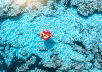 Aerial drone view of woman relaxing on pink inflatable ring in clear turquoise sea above rocks. Summer vacation, Mallorca, Balearic Islands, Spain. Top down view of swimming girl, blue water. Travel © den-belitsky