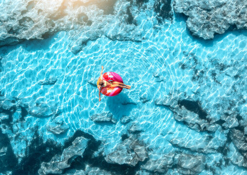 Aerial drone view of woman relaxing on pink inflatable ring in clear turquoise sea above rocks. Summer vacation, Mallorca, Balearic Islands, Spain. Top down view of swimming girl, blue water. Travel
