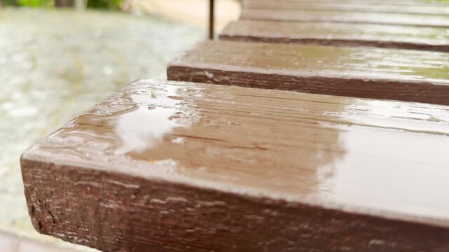 Rainwater pools on the corner of a brown wooden bench during a gentle spring shower. The shiny wet slats reflect light next to a blurry stone path.