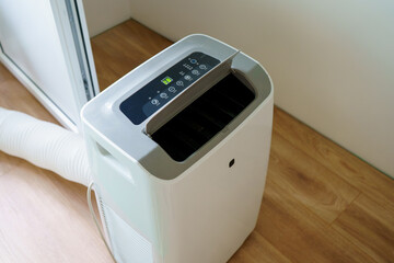 A modern portable mobile air conditioner unit stands on a wooden floor during a hot summer day, controlling temperature and relative humidity inside the room. © Alina Mosinyan