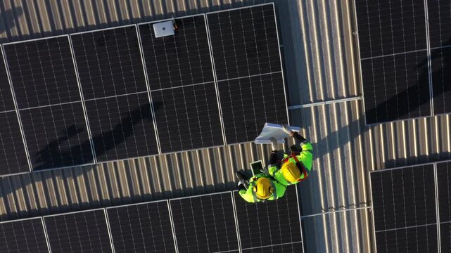 Two workers install solar panels on dark metal rooftop during daylight, wearing yellow hard hats and safety vests to promote renewable energy solutions with professional equipment under focused lighti