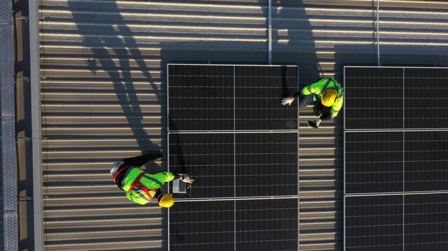 Yellow and green high-visibility workers install solar panels on dark building exterior during an industrial installation project with long shadows highlighting renewable energy technology.