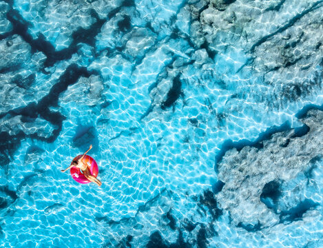 Aerial drone view of woman relaxing on pink inflatable ring in clear turquoise sea above rocks. Summer vacation, Mallorca, Balearic Islands, Spain. Top down view of swimming girl, blue water. Travel