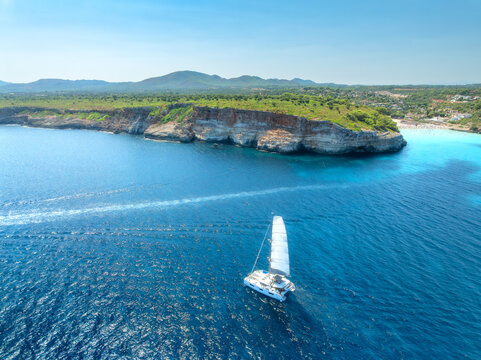 Aerial view of sailing catamaran yacht in turquoise sea water bay. Sailboat. Mallorca, Spain. Luxury boat cruising in ocean cove near sandy beach, resort houses on rocky coast. Drone summer landscape
