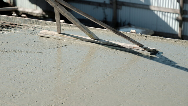 Wet Concrete Slab Leveled With Wooden Screed. Wet concrete slab being leveled with a wooden screed on a sunlit construction site, with formwork edges and metal siding blurred in the background.