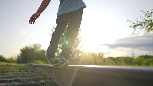 Boy walking on railway track balancing on rail. Child legs in sneakers step on sunny railroad. Kid walks safely in warm summer sunset. Little boy plays on train track outdoors in green nature.