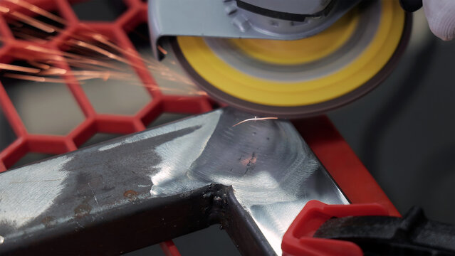 Angle Grinder Smoothing Welded Metal Frame. Close-up angle grinder smooths a welded steel frame, sending sparks across a red clamp fixture in a workshop setting.