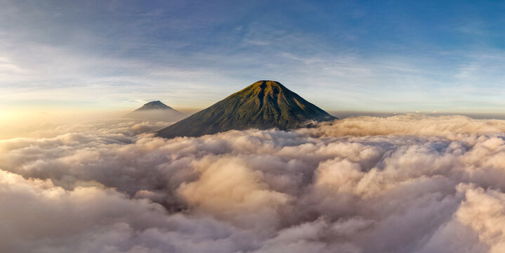 Aerial view of the majestic Mount Sindoro and Mount Sumbing rising above a dense sea of clouds during golden hour in Kecamatan Kedu, Jawa Tengah, Indonesia.