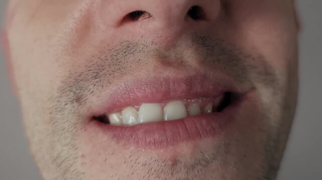 Macro shot captures an unshaven male lower face giving a slight subtle smile. The expression shows upper teeth and the texture of lips and stubble against a plain gray background.