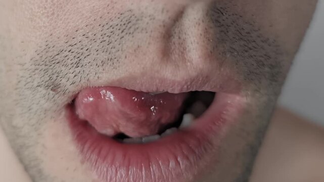 Extreme close up of unshaven man licking dry lips with tongue. Detailed view of male mouth with stubble moving tongue over lips. Concept represents hunger, thirst, or anticipation.