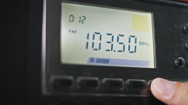 Close-up of a finger holding an auto scan button on a digital radio tuning down the FM band. The LCD screen shows changing MHz numbers and a signal meter on the black receiver panel.