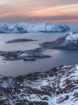 Aerial view of the town of Unalaska nestled among snow-covered mountains and icy waters under a soft sunset sky in Unalaska, Alaska, United States.