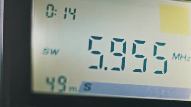 Macro view of a digital radio LCD screen scanning longwave and shortwave bands. Teal numbers change rapidly showing kHz and MHz frequencies while searching for a broadcast signal.