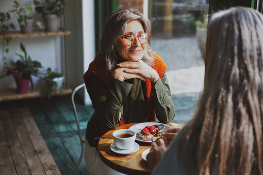 A grey haired woman in an orange scarf, rests her chin on folded hands and smiles softly, while listening to a companion seated across a round wooden table inside a plant filled cafe.
