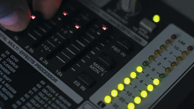 Close-up of a sound engineer's hand adjusting illuminated EQ faders on a digital multi-effects processor. Yellow LED level meters are active in the dark, professional recording studio.