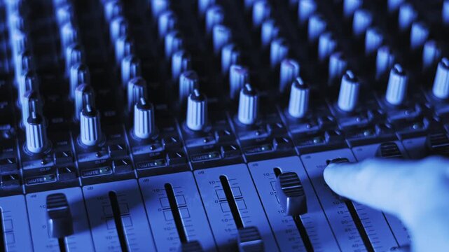 Close-up of a producer adjusting channel faders on a pro audio mixer console. Illuminated mute buttons and knobs glow under dynamic pink and orange lighting in a dark broadcast studio.