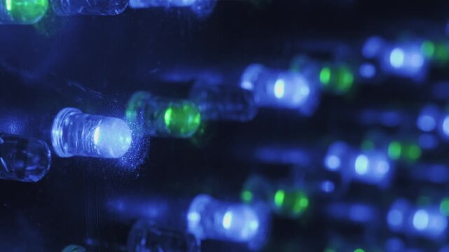 Angled macro shot capturing rows of flashing RGB LED lights on a panel. Shallow depth of field focuses on foreground diodes, creating a dynamic colorful bokeh effect in a dark environment.