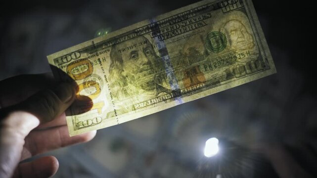Hand reveals the security watermark on a one hundred dollar banknote using a bright LED flashlight in the dark. Concept of detecting counterfeit currency and verifying authentic money.