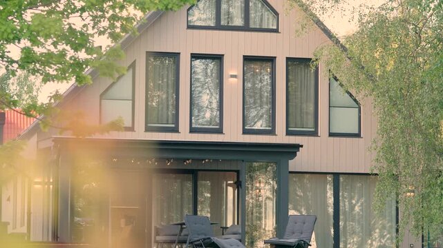Modern wooden house exterior with large windows, green lawn, and outdoor lounge chairs in a serene garden setting