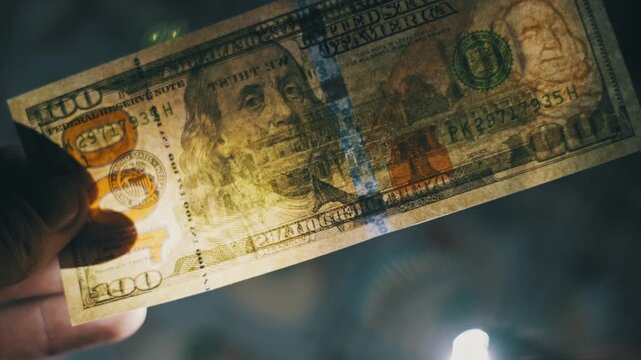Close-up of a hand holding a US one hundred dollar bill against a bright light source to reveal the watermark. The dark moody background highlights the authenticity check.