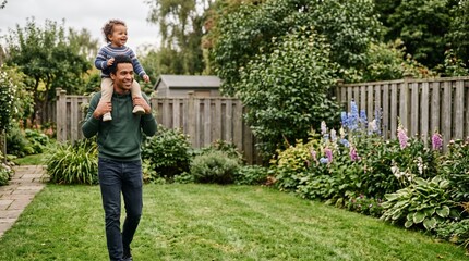 Fototapeta premium Father carrying his happy child on shoulders walking in a lush green backyard garden with flowers