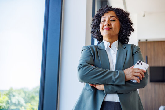 Confident mature businesswoman with curly hair holding mobile phone, arms crossed, standing in office new future