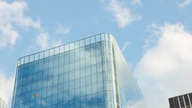 Low-angle architectural view of the top sections of a modern glass office skyscraper with a rounded corner, reflecting a bright blue sky filled with white clouds.