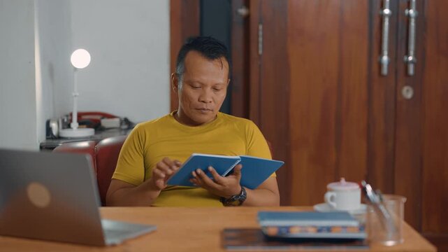 Casual and relaxed Asian man reading a notebook in home office desk. Learning, analyzing and education concept.