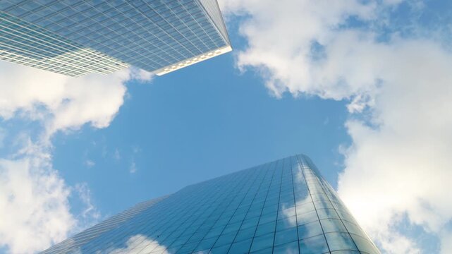 Low-angle architectural view looking straight up into the sky between modern glass skyscrapers, showcasing structural grid patterns and reflections of passing clouds.