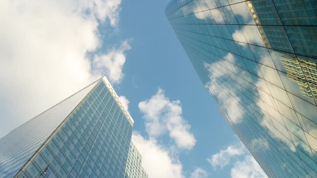 Low-angle architectural view looking up between modern corporate skyscrapers with glass facades, showcasing dramatic reflections of the blue sky and white clouds.