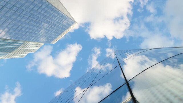 Low-angle architectural view looking straight up into the sky between modern glass skyscrapers, capturing the geometric patterns of the facades and bright reflections of passing clouds.