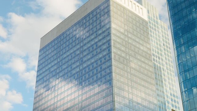 Low-angle architectural view of modern glass office skyscrapers in a commercial business district, with reflections of a bright blue sky and clouds on the glass facades.